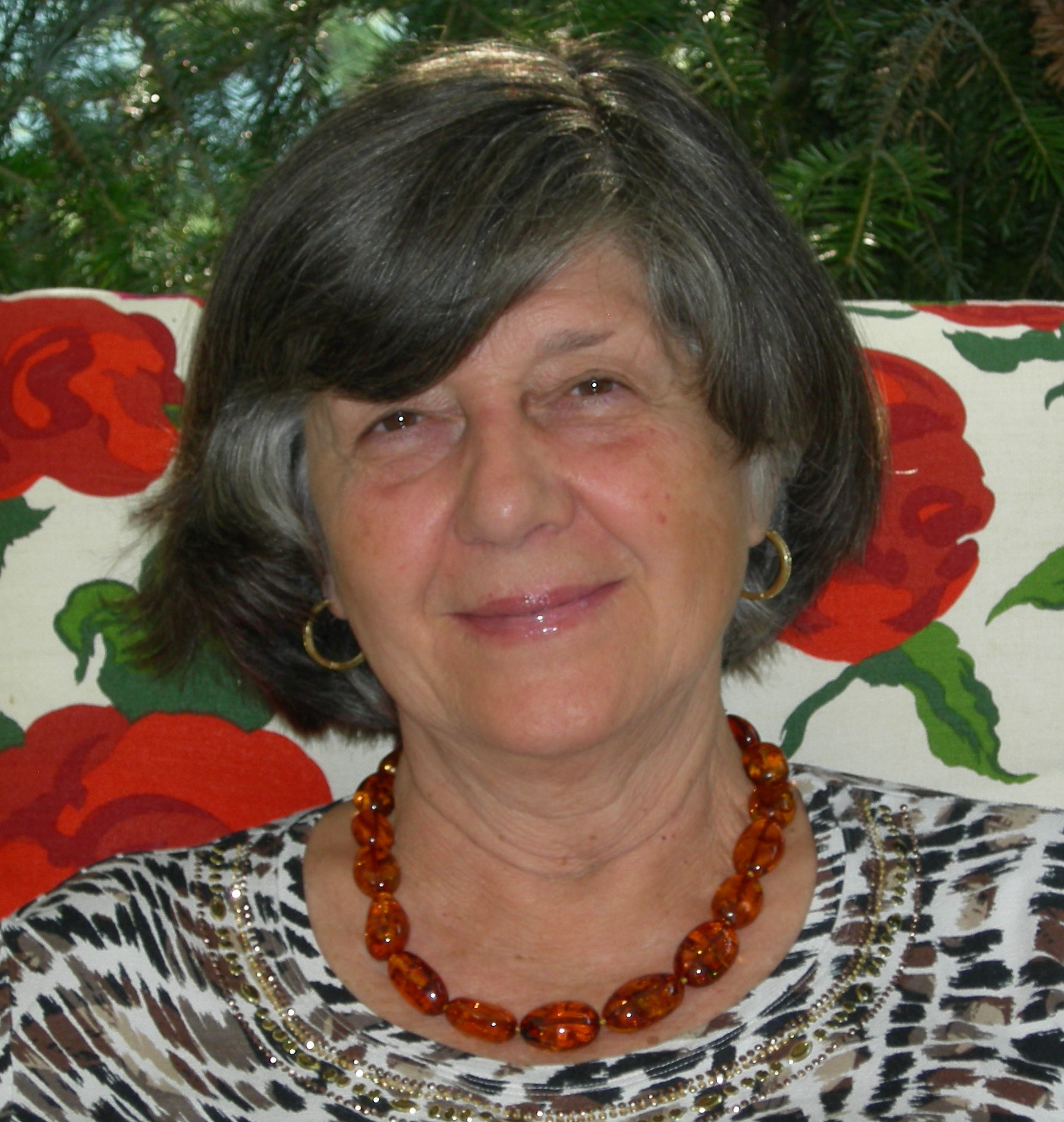 image of Anne Grazia Laura, smiling older white woman with greying hair seated on chair with pattern of large red flowers