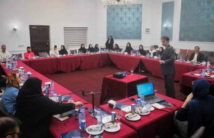 Seminar in a large room with woven tapestries on the walls, tables covered in red tablecloths, forming a large, open square. A speaker is standing on the right, next to a video projector, and about 20 people are sitting at the tables. 