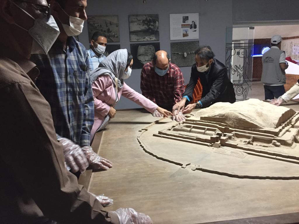 A group of blind tourists around a scale model of an ancient ziggurat, touching parts of the model